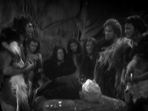 A group of primitive cave people dressed in animal skins stand in a semi-circle around the Doctor, who lies unconscious on a rock.